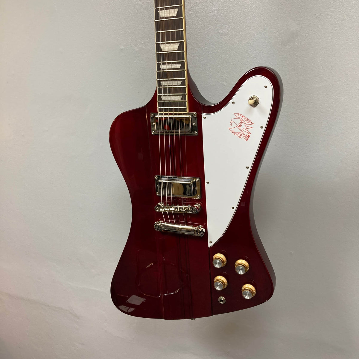 An Epiphone Firebird Wine Red electric guitar with a rosewood fretboard, white pickguard, gold hardware, and three control knobs hangs against a plain light-colored wall. Includes gigbag.