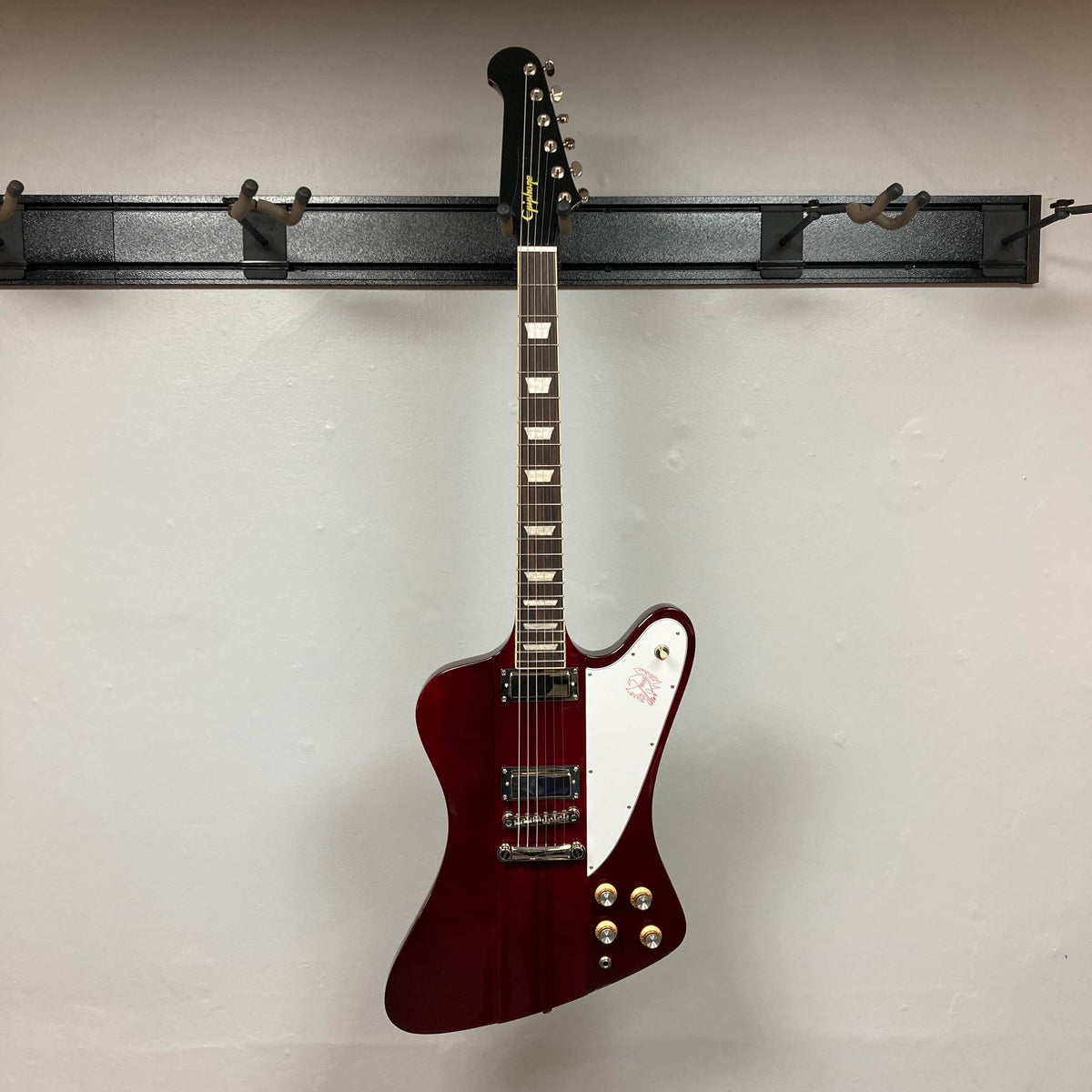 An Epiphone Firebird Wine Red electric guitar with a unique offset body and ProBucker FB720 pickups hangs on a wall rack against a plain white background.