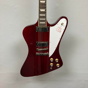 An Epiphone Firebird Wine Red electric guitar with a white pickguard, gold knobs, angular body, and ProBucker FB720 pickups hangs against a light-colored wall. Includes gigbag.