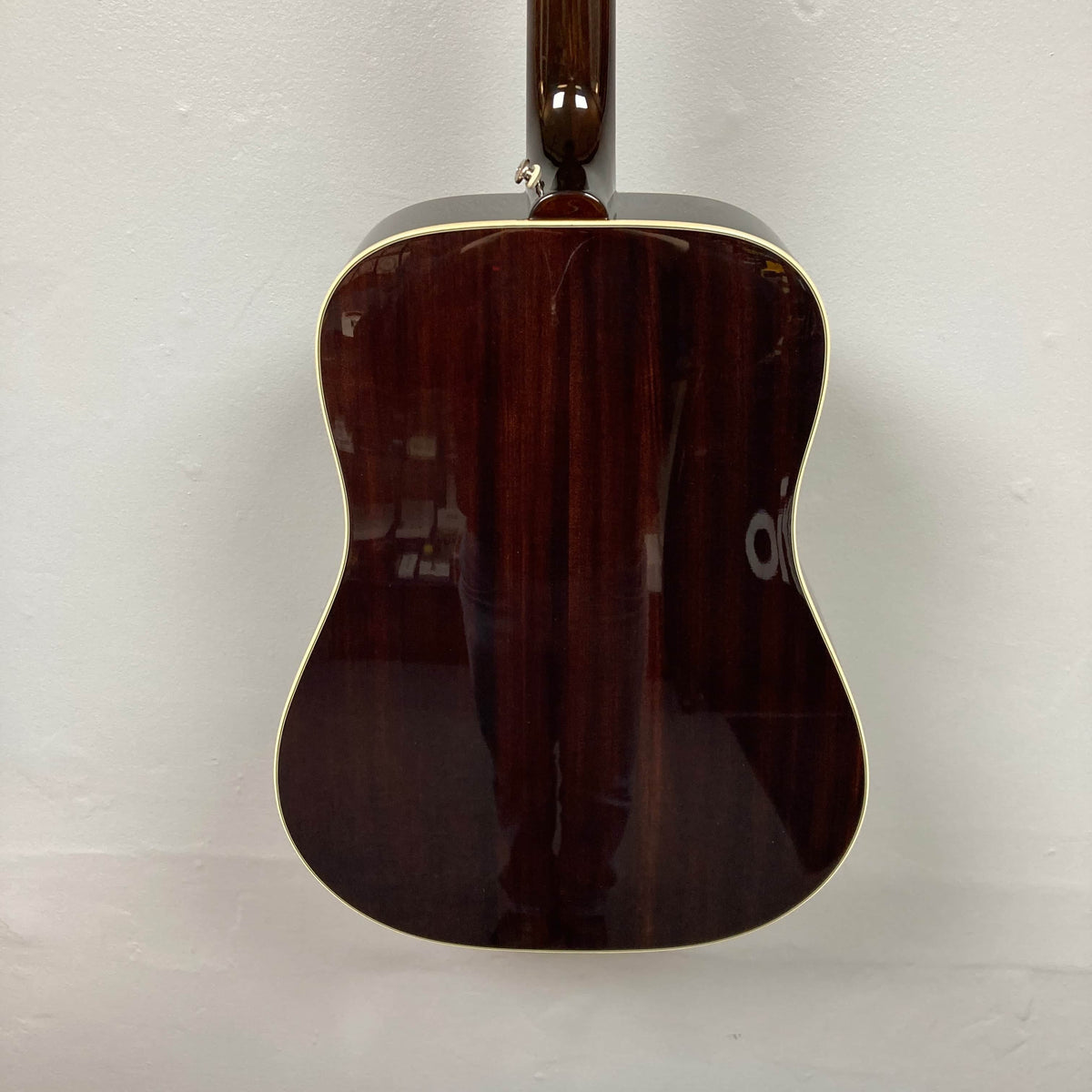 A rear view of the Epiphone Hummingbird Studio Antique Natural w/Gigbag by Epiphone displays its glossy dark wood back with cream binding, photographed against a plain white wall.