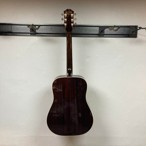An Epiphone Hummingbird Studio Antique Natural acoustic guitar with a solid spruce top is displayed from the back, hanging centrally on a black wall rack against a plain white background. Includes gigbag.