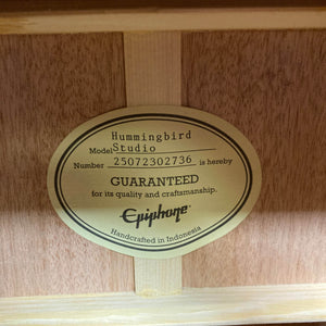 A close-up of a label inside an Epiphone Hummingbird Studio Antique Natural w/Gigbag states: "Model Number 25072302736 is GUARANTEED for quality and craftsmanship. Solid spruce top, handcrafted in Indonesia.