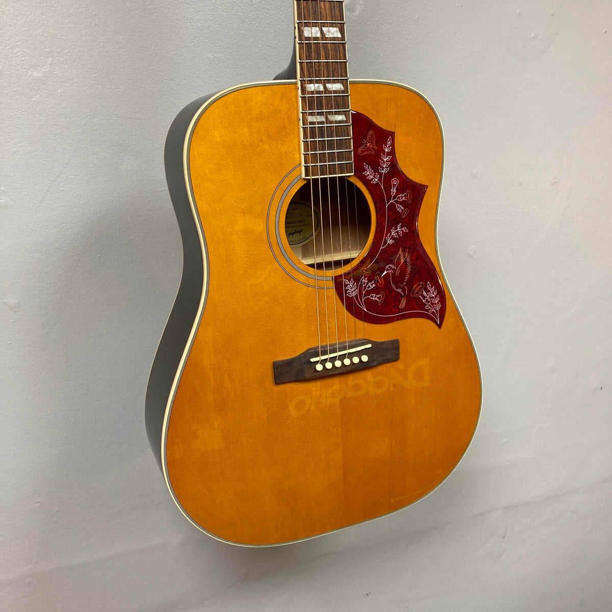 An Epiphone Hummingbird Studio Antique Natural acoustic guitar by Epiphone hangs against a light gray wall, showing its solid spruce top, reddish floral pickguard, white binding, and soundhole. Comes with a gigbag.