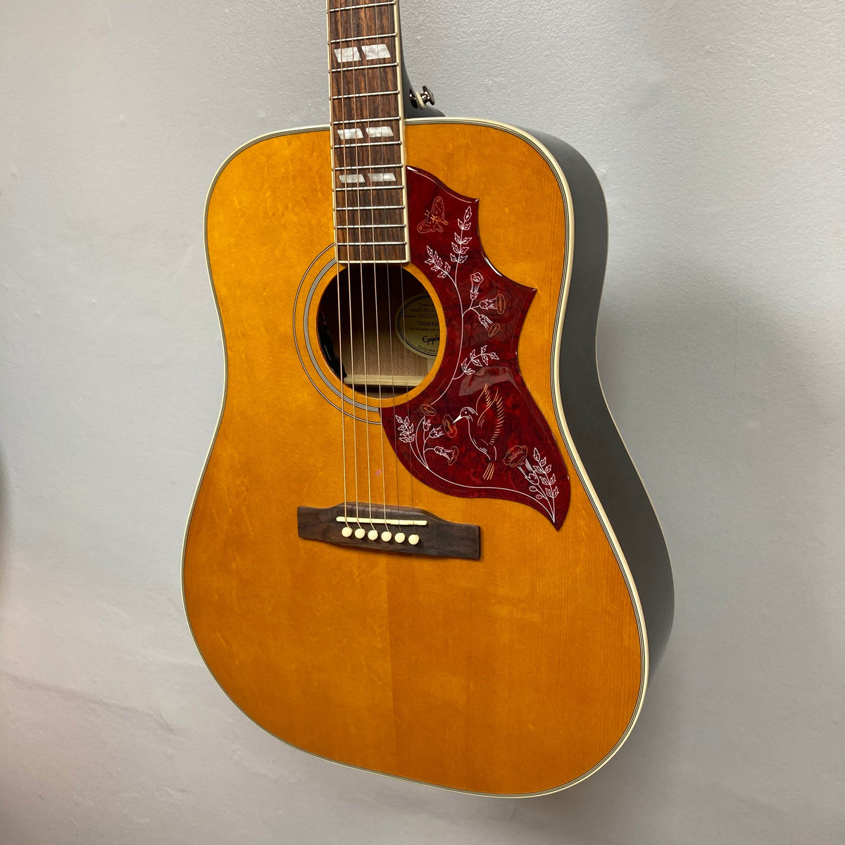 A close-up of the Epiphone Hummingbird Studio Antique Natural acoustic guitar, featuring a solid spruce top, red floral pickguard, six strings, and shown hanging against a plain light wall. Includes gigbag. Brand: Epiphone.