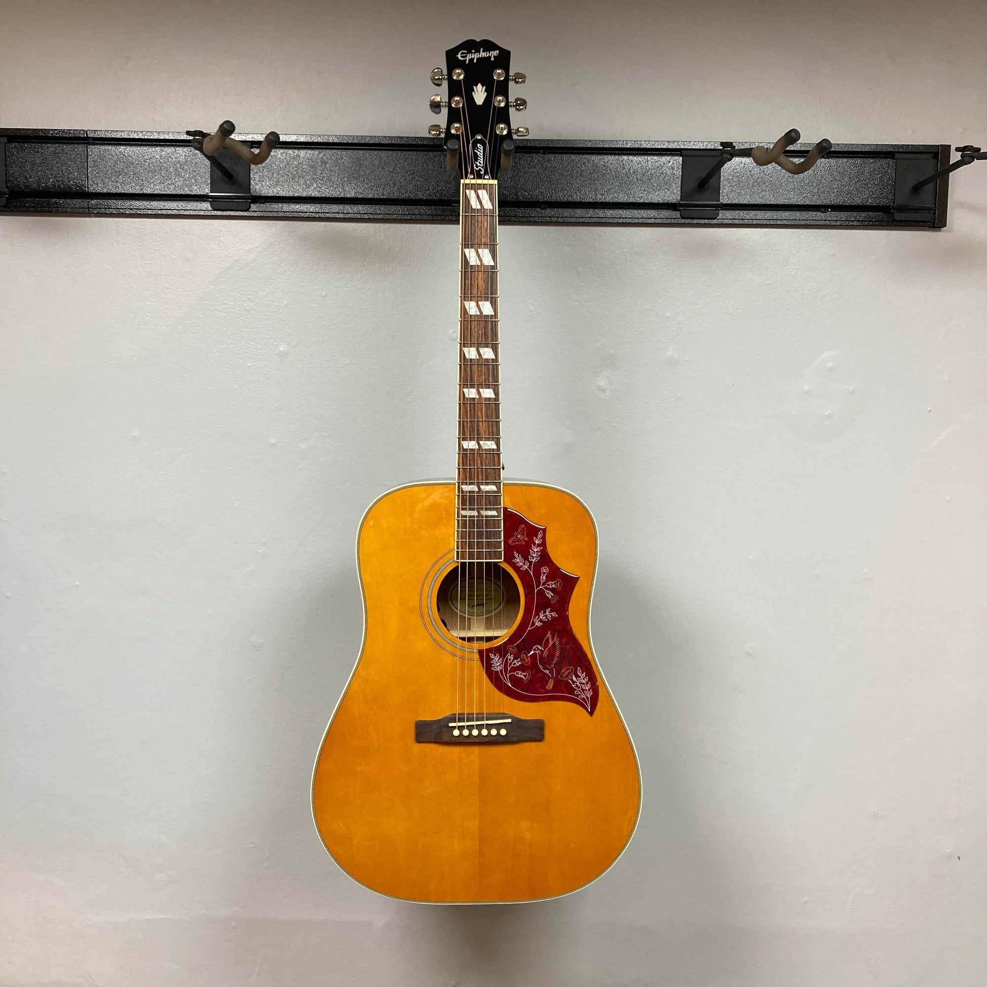An Epiphone Hummingbird Studio Antique Natural acoustic guitar with floral and bird motif on the pickguard, six strings, white fret markers, and solid spruce top, is shown against a plain light gray background.