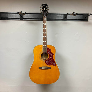 An Epiphone Hummingbird Studio Antique Natural w/Gigbag by Epiphone hangs on a black wall mount, featuring a solid spruce top, red decorative pickguard, mother of pearl block inlays, and a Fishman Sonitone pickup.