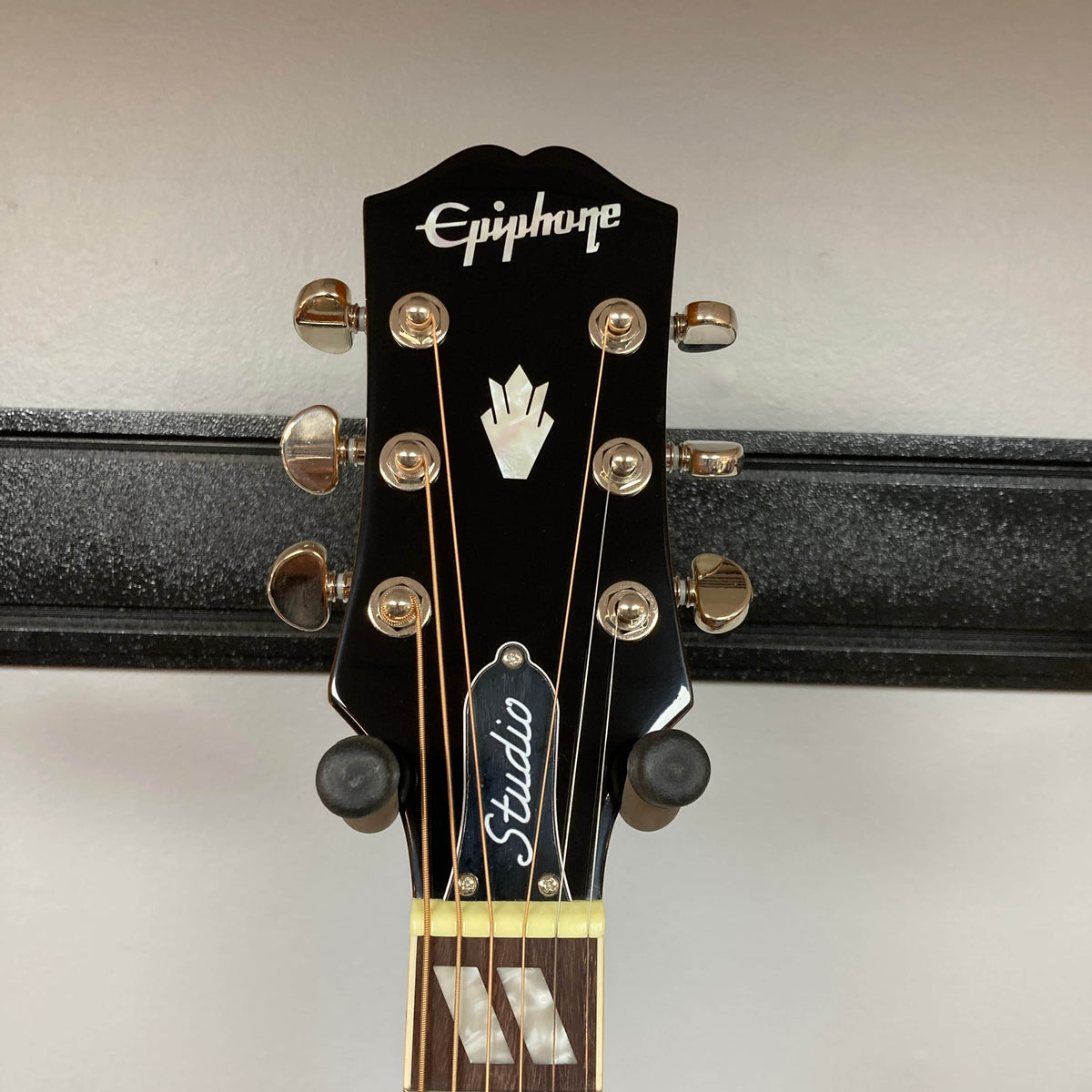 Close-up of the headstock of the Epiphone Hummingbird Studio Faded Cherry Sunburst acoustic-electric guitar, featuring gold tuners, Epiphone logo, decorative inlay, and "Studio" truss rod cover against a plain light wall.