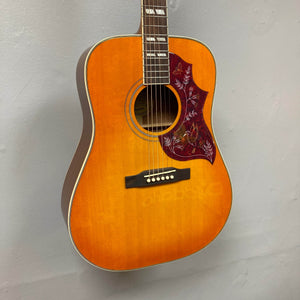 An Epiphone Hummingbird Studio Faded Cherry Sunburst acoustic-electric guitar with Fishman Sonitone pickup, floral pickguard, and white double fretboard inlays hangs on a light gray wall. Brand: Epiphone. Includes gigbag.