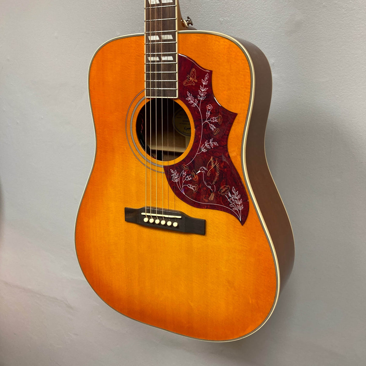 A close-up of the Epiphone Hummingbird Studio Faded Cherry Sunburst acoustic-electric guitar, featuring a decorative pickguard and Fishman Sonitone pickup, hanging on a light gray wall. Includes Epiphone gigbag.