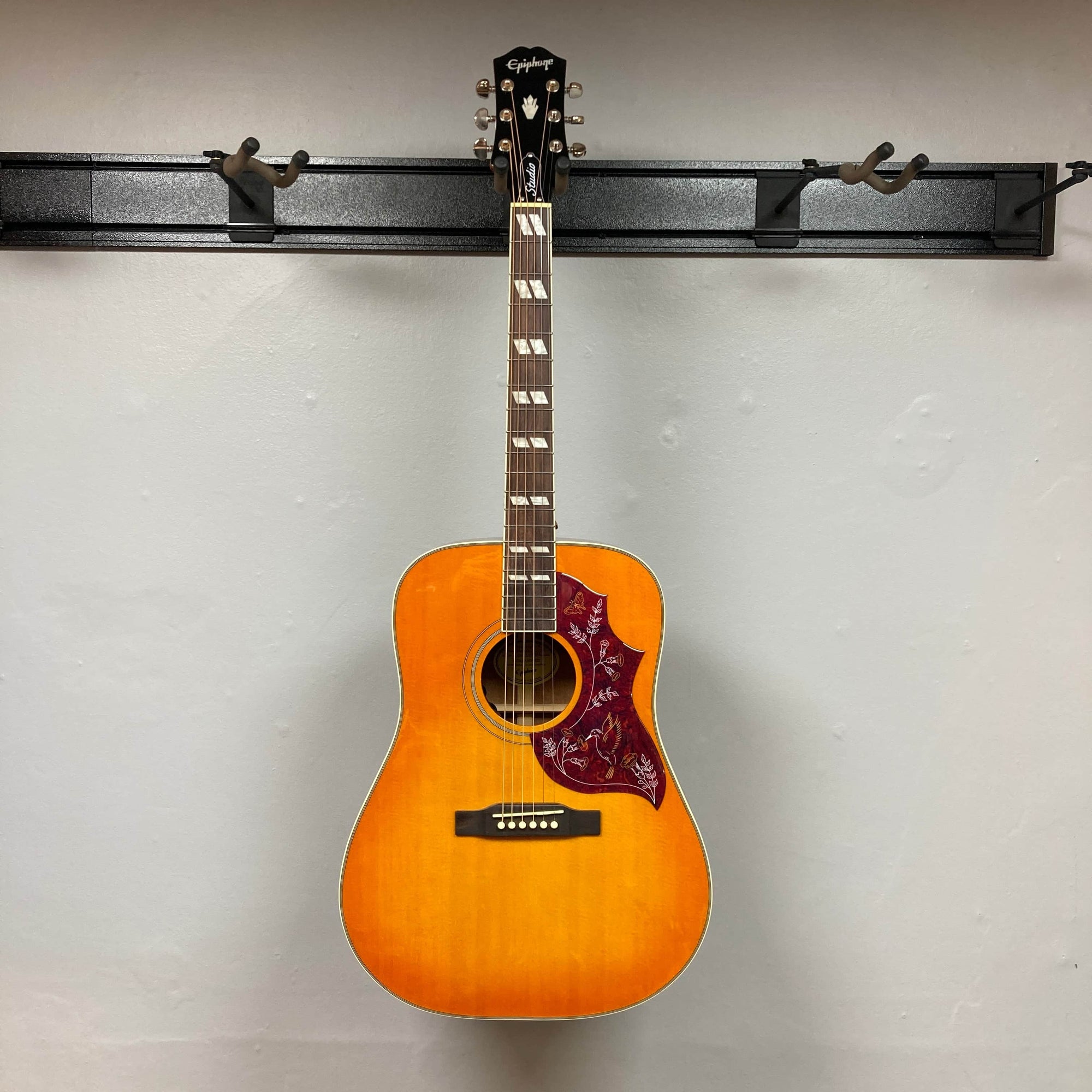 An Epiphone Hummingbird Studio Faded Cherry Sunburst acoustic-electric guitar by Epiphone, featuring a floral red pickguard, six strings, fretboard inlays, Fishman Sonitone pickup, and photographed against a plain light gray wall.