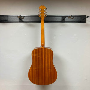 An Epiphone Hummingbird Studio Faded Cherry Sunburst w/Gigbag hangs on a black wall-mounted rack, shown from the back, highlighting its glossy finish and silver tuning pegs against a plain white wall.