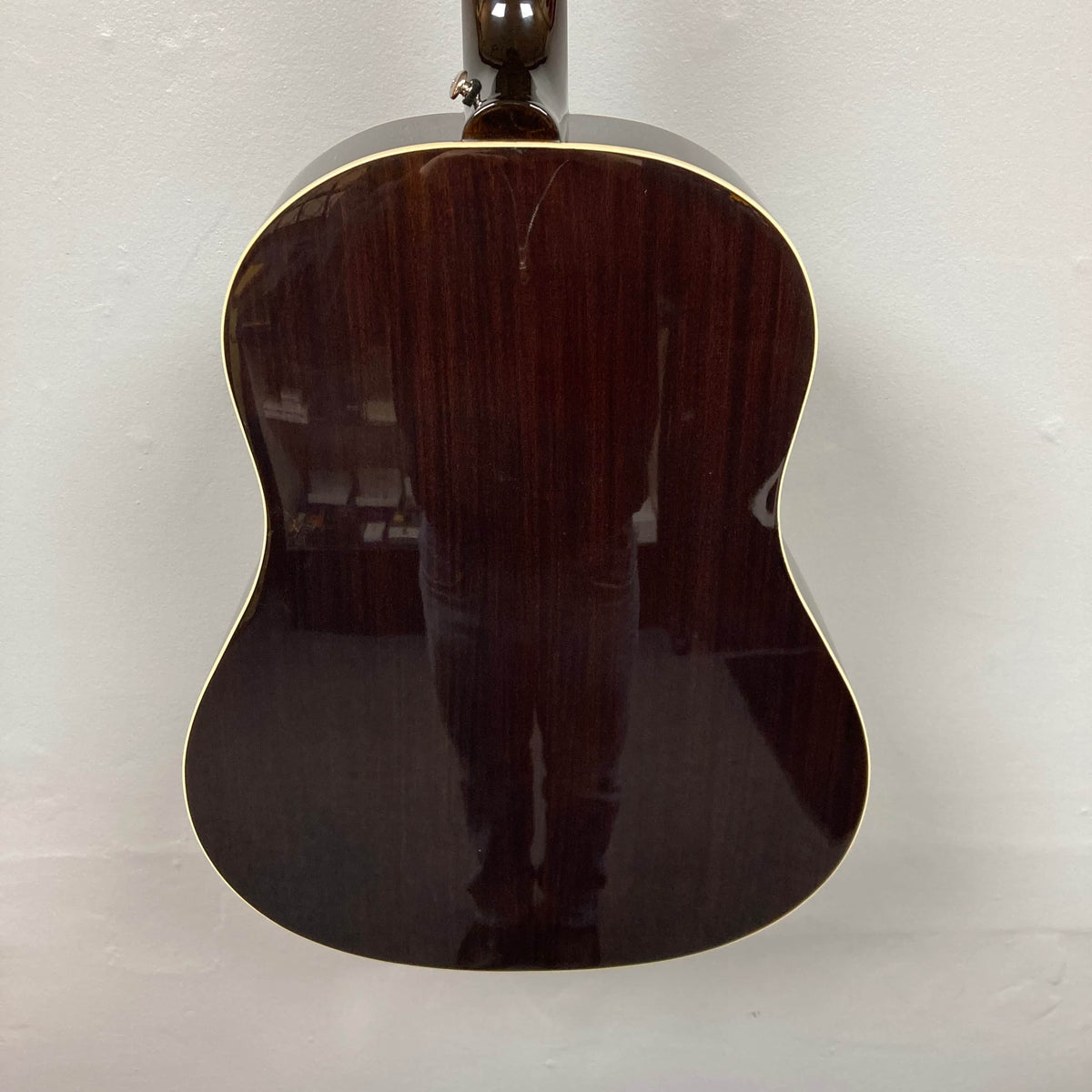 The image shows the back of an Epiphone J-45 Studio Vintage Sunburst acoustic guitar by Epiphone, featuring a dark glossy finish and cream binding. The guitar’s surface reflects a person’s legs. Includes gigbag.