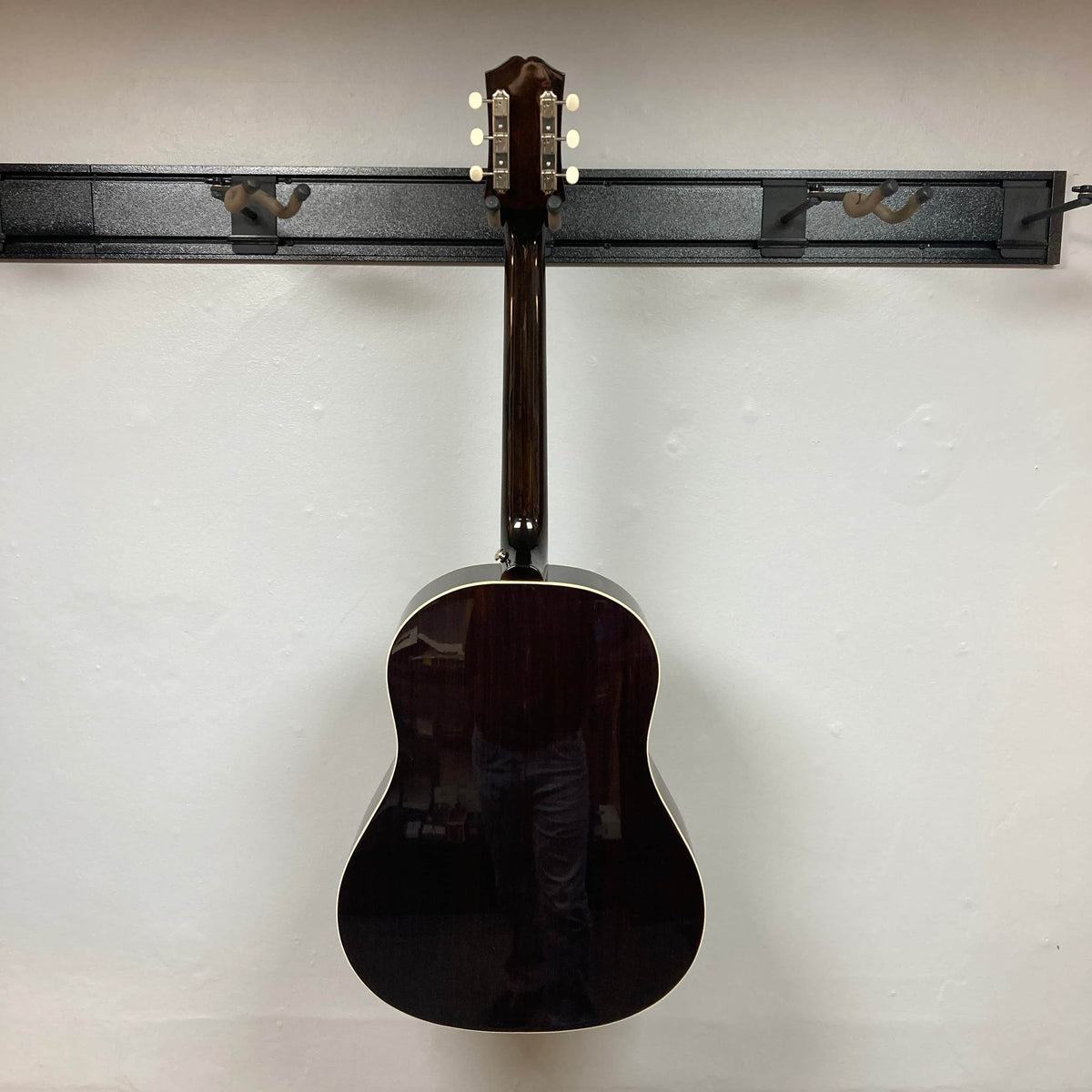 An Epiphone J-45 Studio Vintage Sunburst acoustic guitar hangs on a wall rack, its glossy dark wood finish, light binding, and solid spruce top standing out against the plain white wall. Includes Gigbag.
