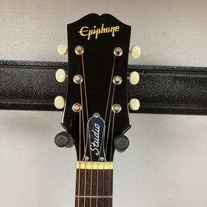 Close-up of the Epiphone J-45 Studio Vintage Sunburst headstock by Epiphone, showing six tuners, gold logo, "Studio" truss rod cover, and fretboard. This round shoulder acoustic guitar with solid spruce top rests on a black wall mount.