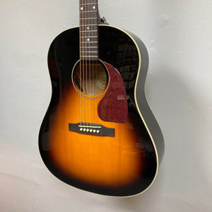 An Epiphone J-45 Studio Vintage Sunburst w/Gigbag acoustic guitar by Epiphone hangs on a plain light wall, its glossy finish, steel strings, and red-brown pickguard highlighting the sound hole and bridge.