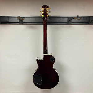An Epiphone Jerry Cantrell Wino Les Paul Custom in Wine Red with gold tuning pegs and white trim, shown from the back, hangs on a wall-mounted rack. Case included.