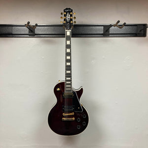 An Epiphone Jerry Cantrell Wino Les Paul Custom electric guitar in wine red with gold hardware and white trim hangs on a wall-mounted rack against a plain white background. Comes with case.
