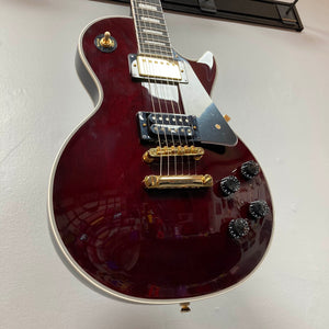A close-up of the Epiphone Jerry Cantrell Wino Les Paul Custom in Wine Red with gold hardware and black accents, hanging on a wall. Includes a custom hardshell case for safe storage and transport.