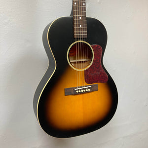 An Epiphone L-00 Standard Vintage Sunburst w/Gigbag, a small-bodied acoustic guitar with a red pickguard, hangs on a light-colored wall.