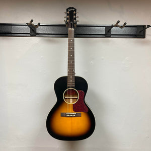 A compact Epiphone L-00 Standard Vintage Sunburst acoustic guitar with gigbag hangs on a black wall-mounted rack against a plain white wall.