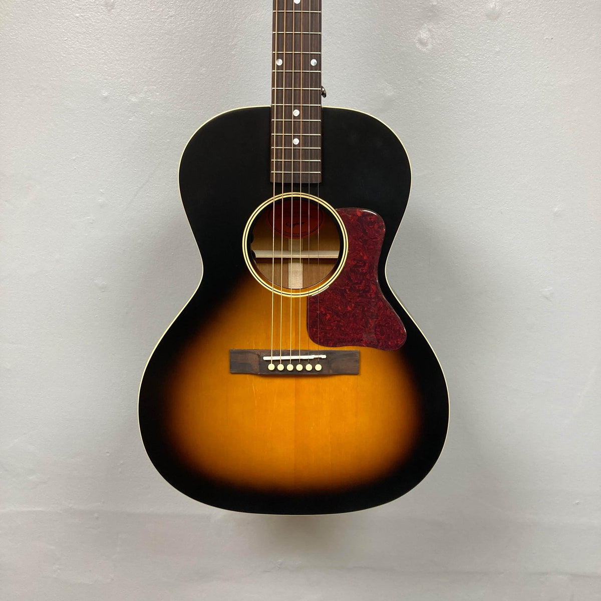 The Epiphone L-00 Standard Vintage Sunburst w/Gigbag, a small-bodied six-string acoustic guitar with a red tortoiseshell pickguard, hangs against a plain light-colored wall. Brand: Epiphone.