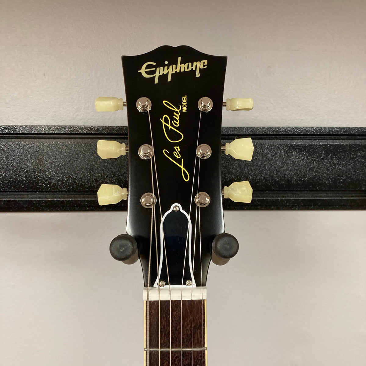 Close-up of an Epiphone Les Paul 1957 Goldtop Reissue Blem headstock, featuring six tuning pegs with white keys and visible logo, as it hangs on a wall-mounted holder. Model comes with case.