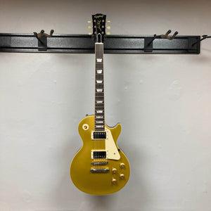 An Epiphone Les Paul 1957 Goldtop Reissue electric guitar, featuring a blemish finish and case, is displayed on a black wall-mounted rack against a white wall.