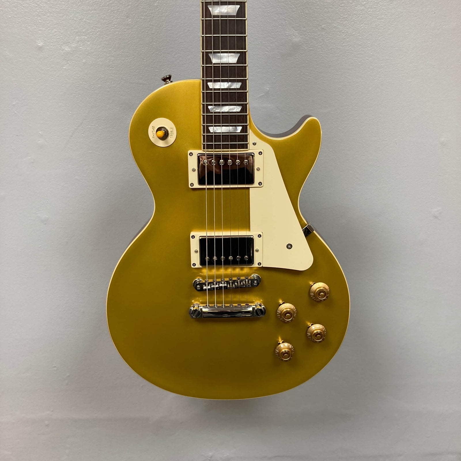 An Epiphone Les Paul 1957 Goldtop Reissue Blem electric guitar with four control knobs, humbucker pickups, a white pickguard, and a single cutaway body hangs against a plain light gray wall. Includes case.