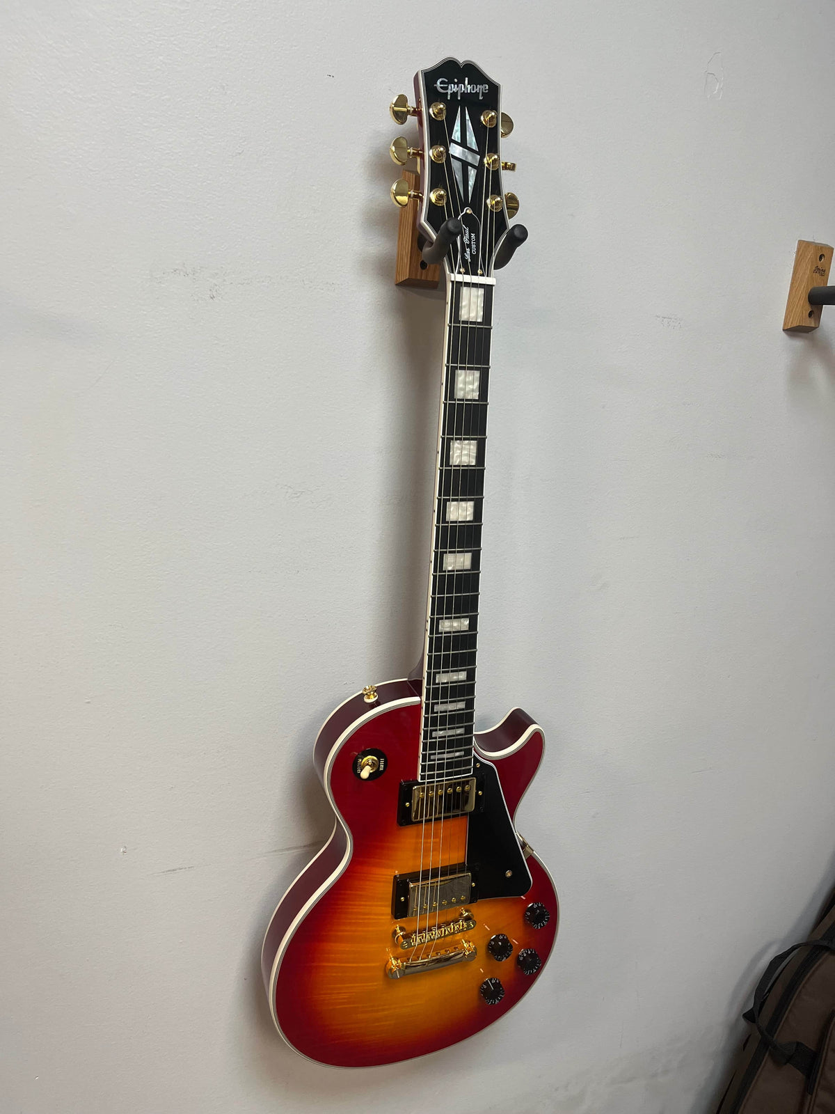 An Epiphone Les Paul Custom, Heritage Cherry Sunburst Exclusive, hangs on a wooden wall-mounted guitar holder.