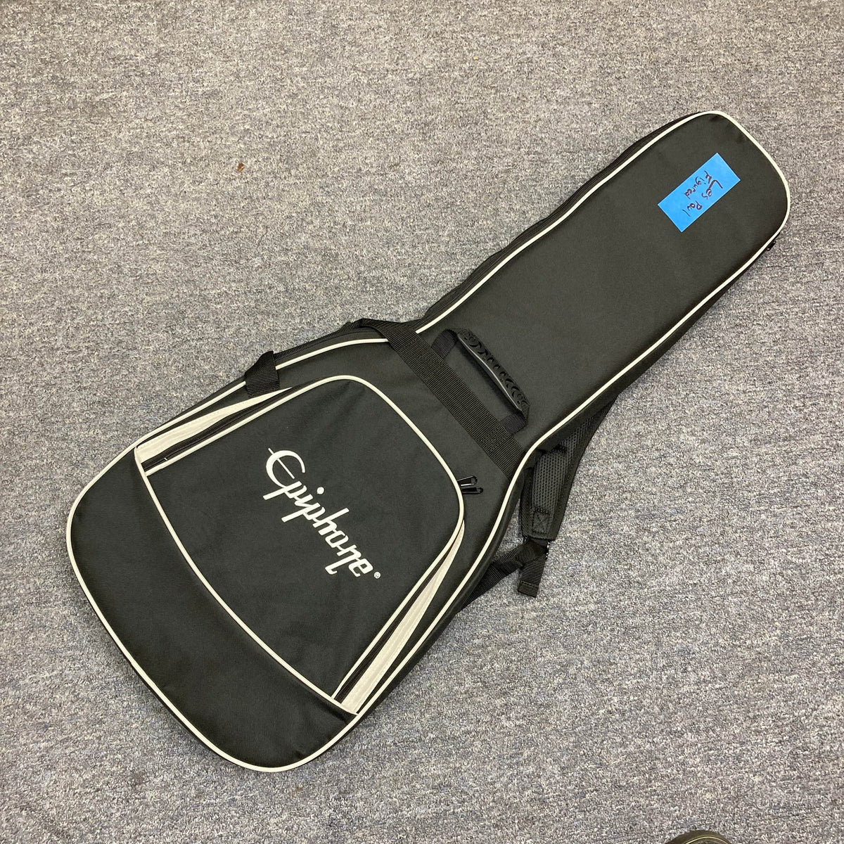 A black Epiphone gig bag with white trim and a front pocket lies on gray carpet, featuring a blue sticky note, ideal for storing your Epiphone Les Paul Custom, Heritage Cherry Sunburst (Exclusive w/GigBag) by Epiphone.