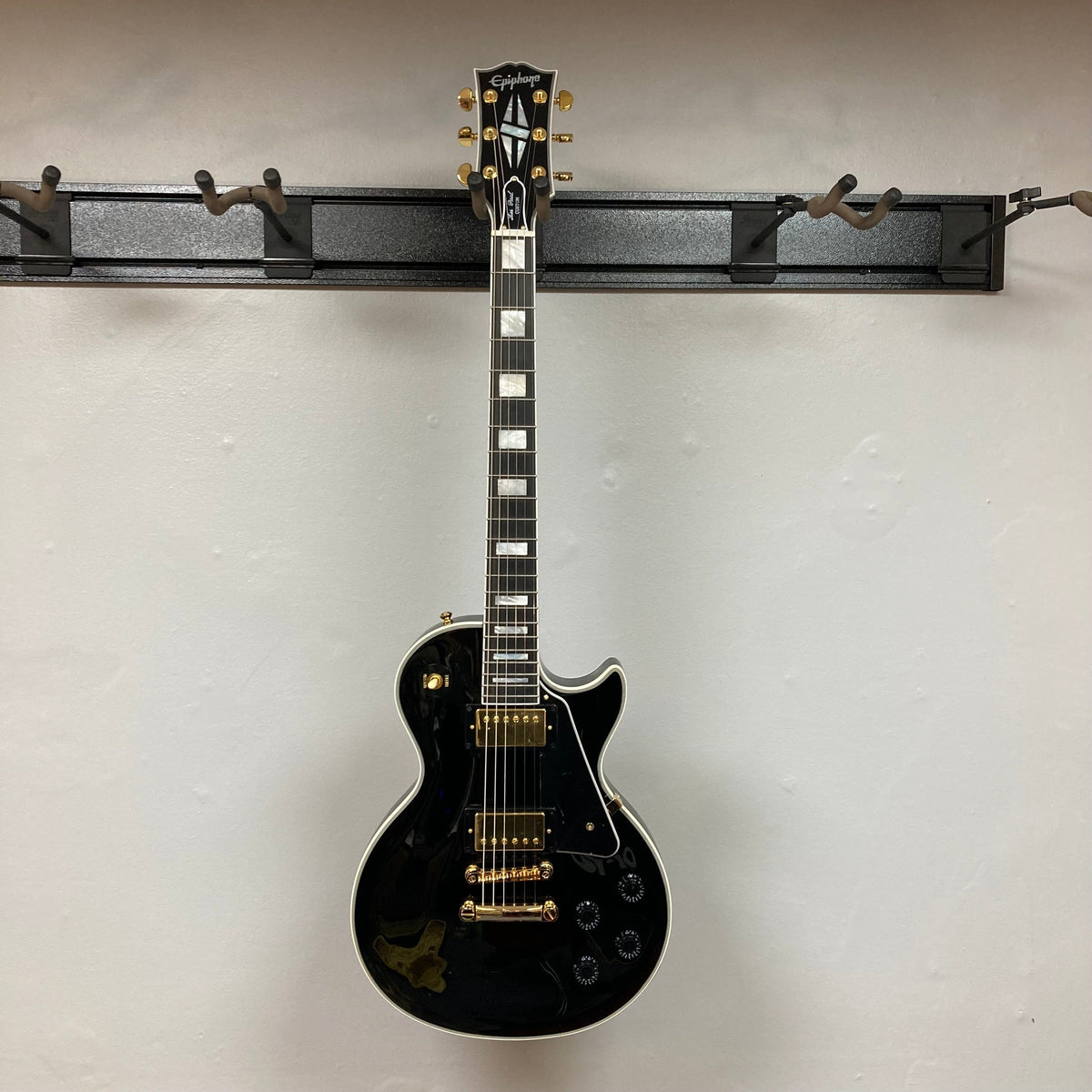 Epiphone Les Paul Custom Inspired by Gibson Ebony w/Case, featuring gold hardware, white squares, and a classic design, hanging on a wall.