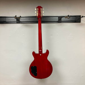 An Epiphone Les Paul Special Double Cut Cherry Red w/Gigbag electric guitar hangs on a wall rack, shown from the back with a black cover plate on its lower left side, echoing Gibson’s iconic design.