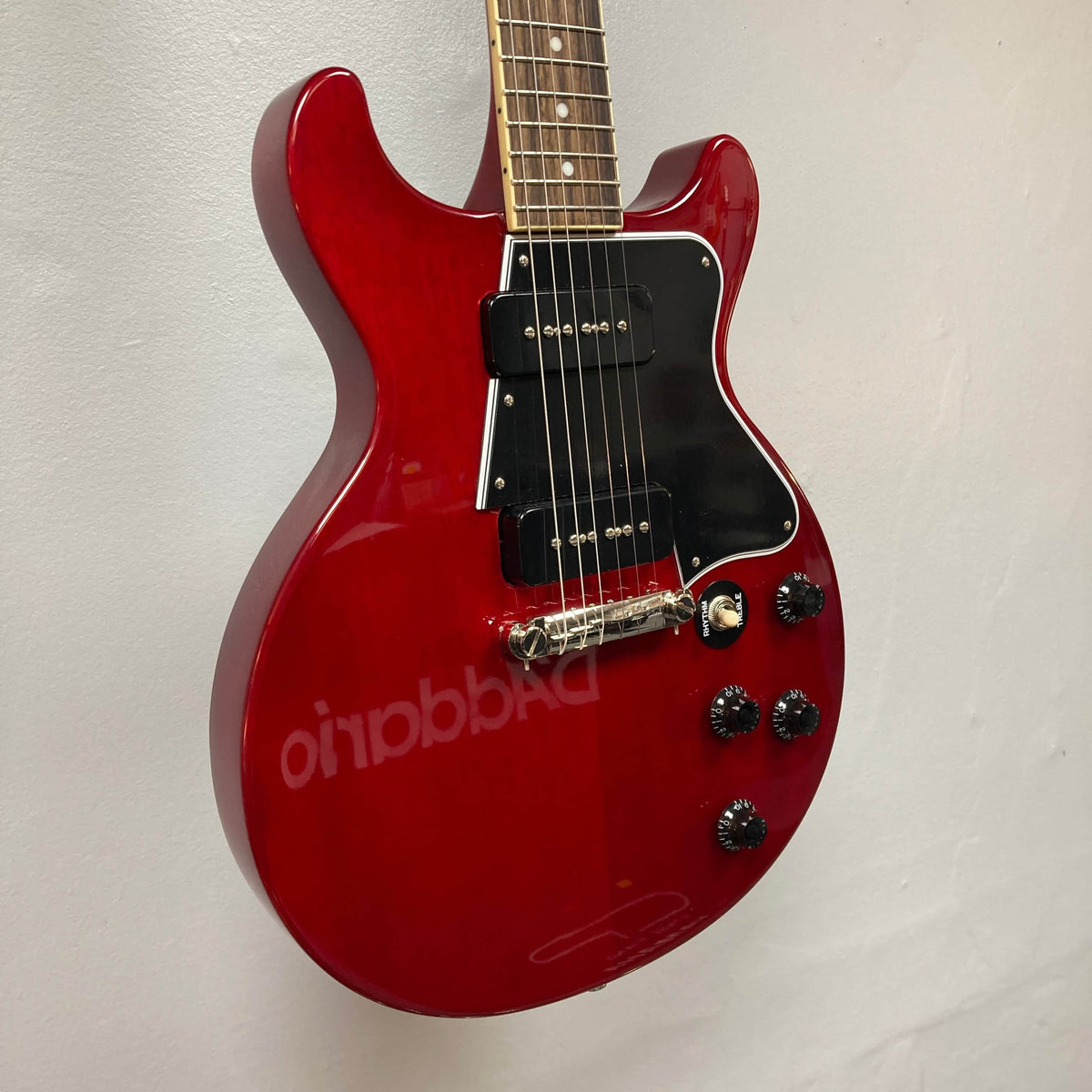 An Epiphone Les Paul Special Double Cut Cherry Red electric guitar with two pickups, four control knobs, and a black pickguard, inspired by Gibson’s 60s SlimTaper neck, is mounted on a white wall. Gigbag included.
