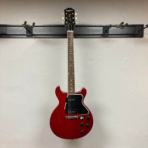 An Epiphone Les Paul Special Double Cut Cherry Red w/Gigbag electric guitar by Epiphone is displayed on a wall-mounted holder against a white background, inspired by Gibson’s classic design.