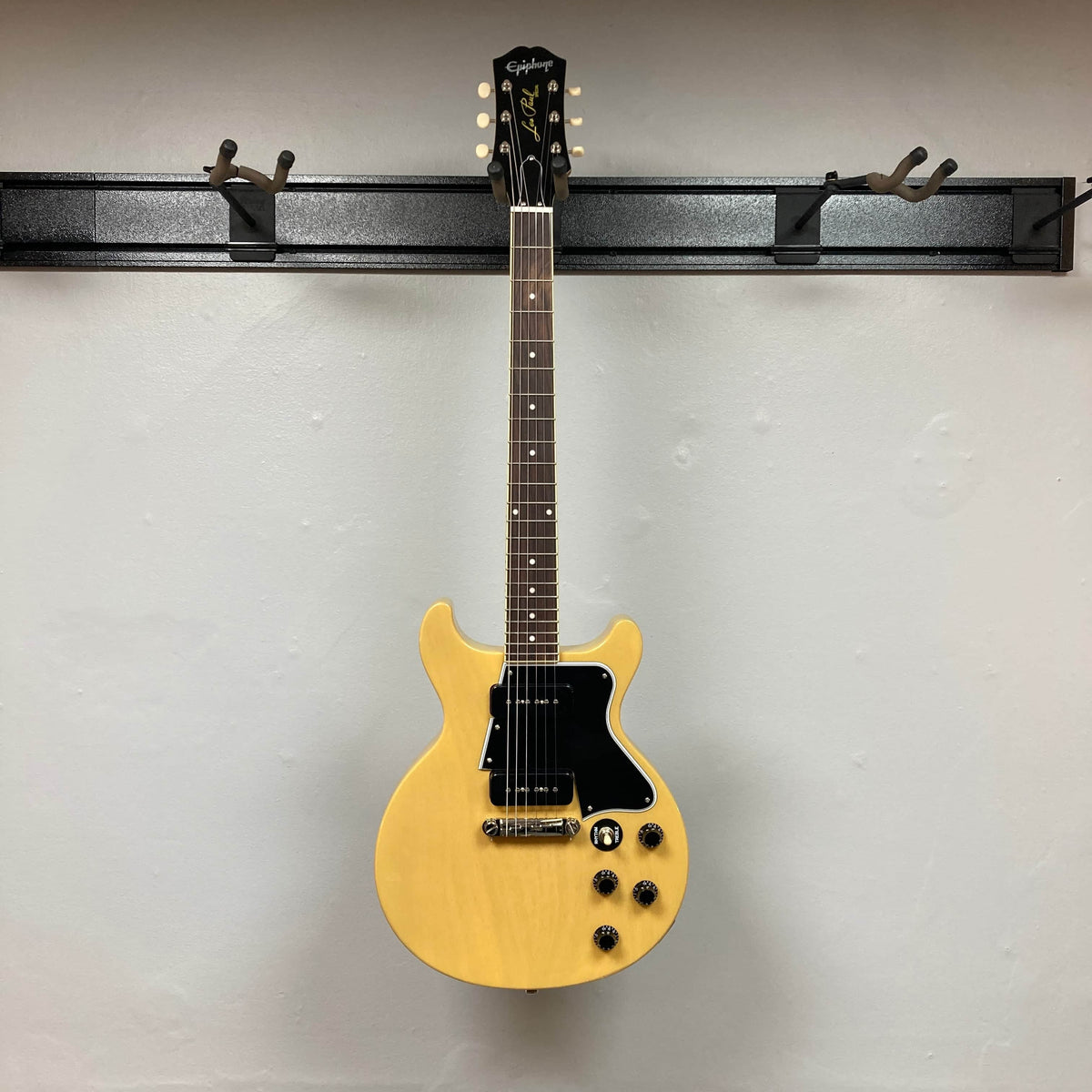 An Epiphone Les Paul Special Double Cut TV Yellow electric guitar with gigbag hangs on a wall-mounted rack against a plain white wall.