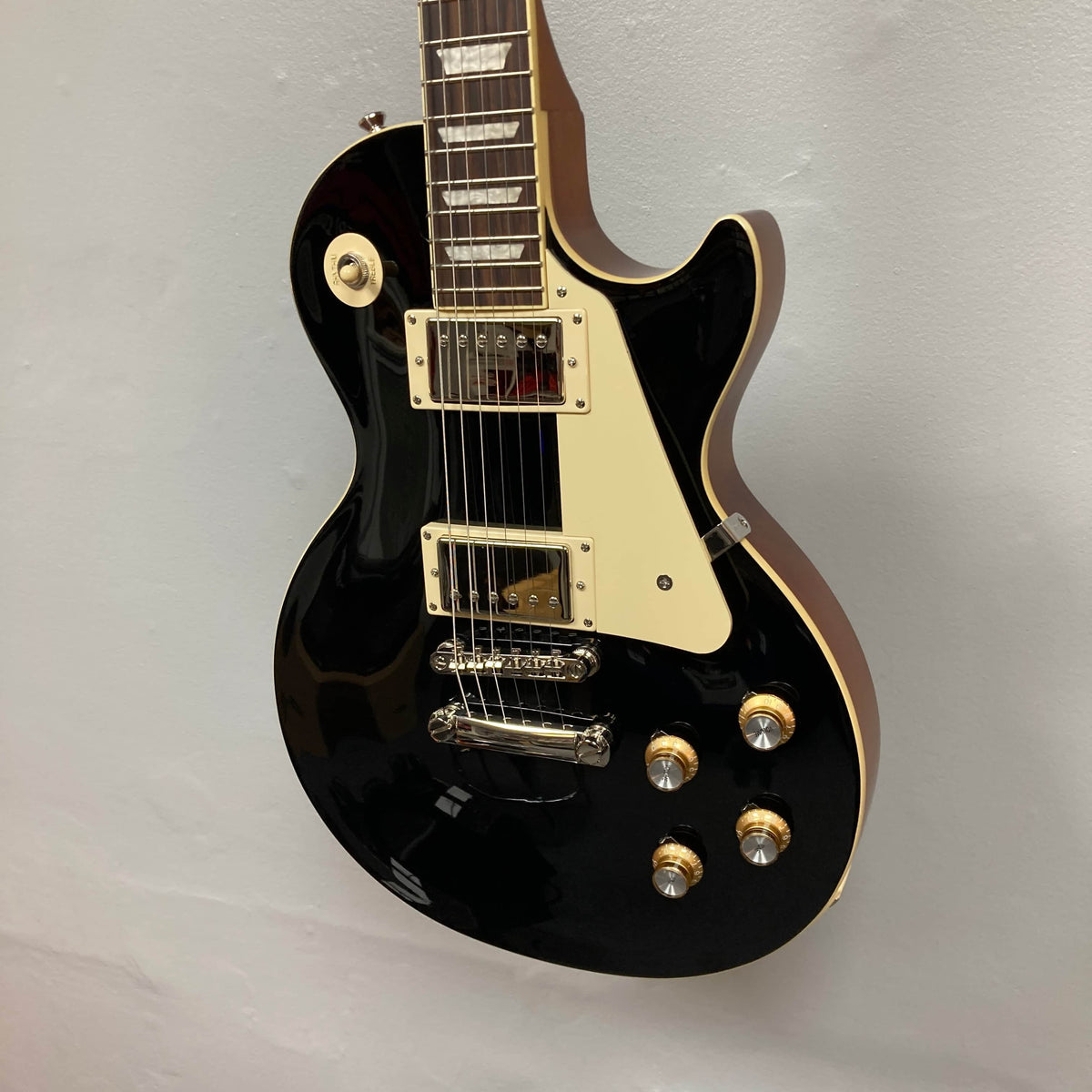 This Epiphone Les Paul Standard 60s Ebony w/Gigbag displays a cream pickguard, gold and silver knobs, and chrome hardware, hanging on a plain light wall.