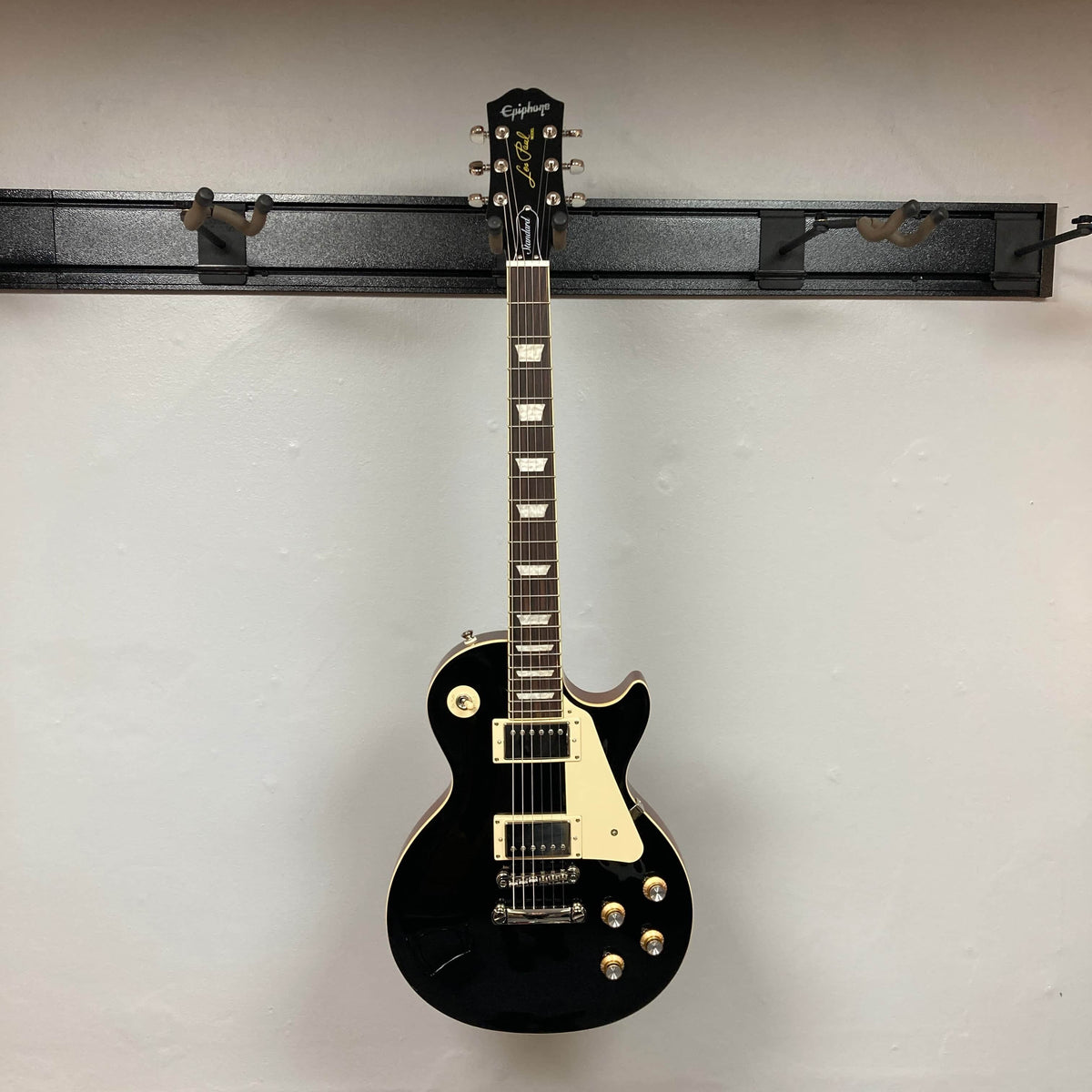 An Epiphone Les Paul Standard 60s Ebony w/Gigbag electric guitar hangs on a wall-mounted rack against a plain, light-colored wall.