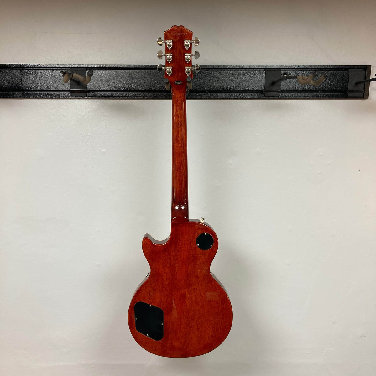 An Epiphone Les Paul Standard 60s Figured Iced Tea Burst electric guitar with visible electronics cavity covers hangs on a wall-mounted rack, shown from the back.
