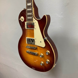 An Epiphone Les Paul Standard 60s Figured Iced Tea Burst electric guitar with humbucker pickups, white pickguard, and gold knobs hangs on a light gray wall.