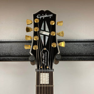 Close-up of an Epiphone Matt Heafy Les Paul Custom Origins, Bone White headstock with gold tuners, mother-of-pearl inlays, and Epiphone logo, mounted on a black wall hanger.