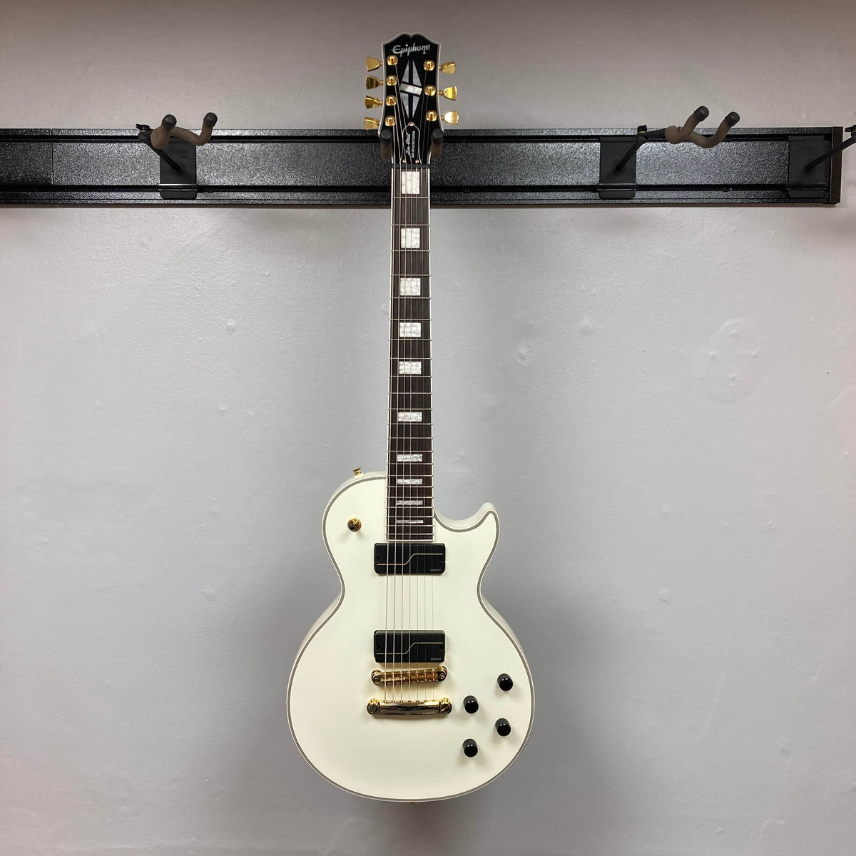 An Epiphone Matt Heafy Les Paul Custom Origins in Bone White with gold hardware, black knobs, and Fishman Fluence pickups hangs on a wall-mounted rack against a plain white background.