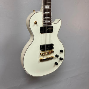 An Epiphone Matt Heafy Les Paul Custom Origins electric guitar in Bone White with black Fishman Fluence pickups, gold hardware, and four black knobs hangs against a light gray wall.