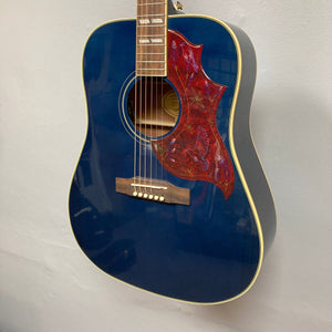 Epiphone Miranda Lambert Bluebird Studio - Bluebonnet guitar featuring red and gold details, hummingbird-style pickguard, and onboard electronics, displayed on a wall.