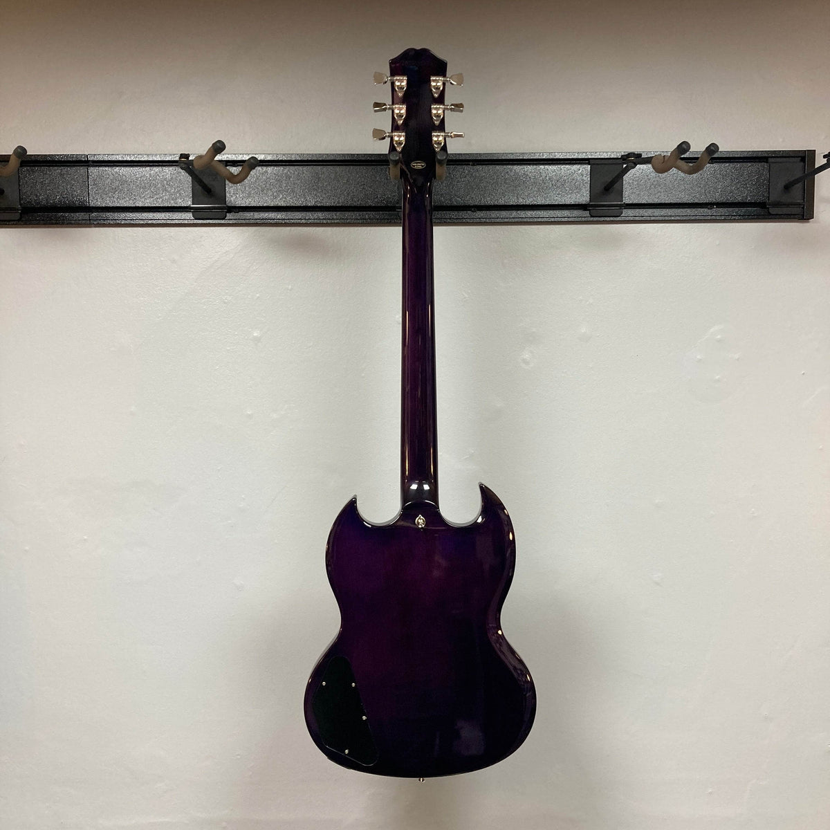 An Epiphone SG Modern Figured Purple Burst w/GigBag electric guitar hangs on a wall rack, shown from the back against an off-white wall. It features a double-cutaway body and silver tuning pegs.