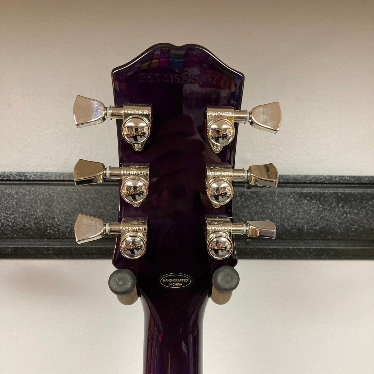 The back of an Epiphone SG Modern Figured Purple Burst w/GigBag headstock shows six tuning pegs, a serial number, and a “Hand Crafted in China” sticker, set against a plain wall.