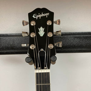The image shows the headstock of an Epiphone SG Modern Figured Purple Burst w/GigBag, featuring six tuning pegs, a decorative inlay, and a truss rod cover, resting on a black stand against a white wall.
