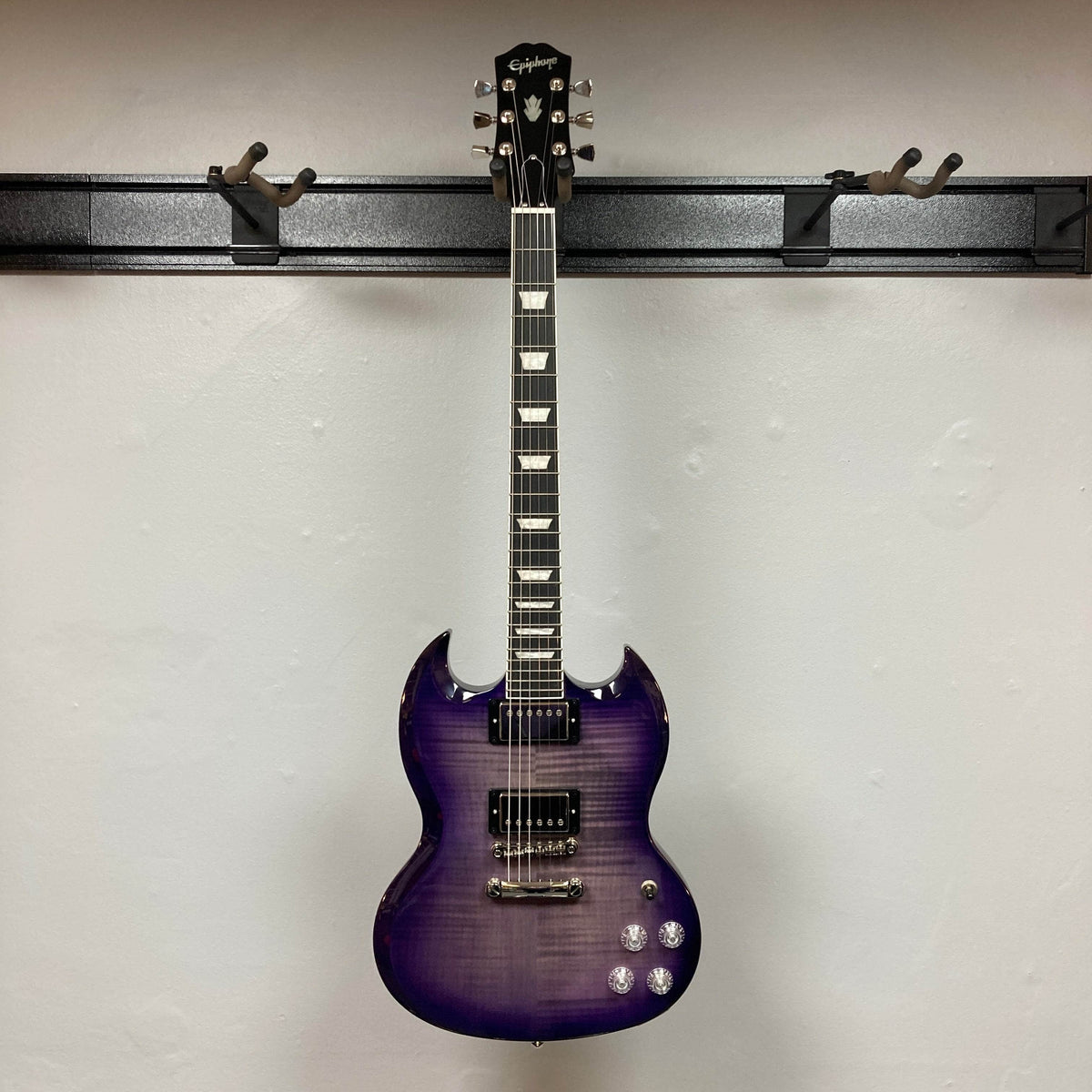 An Epiphone SG Modern Figured Purple Burst electric guitar with chrome hardware hangs on a black wall-mounted holder against a white wall. Included is a gig bag.