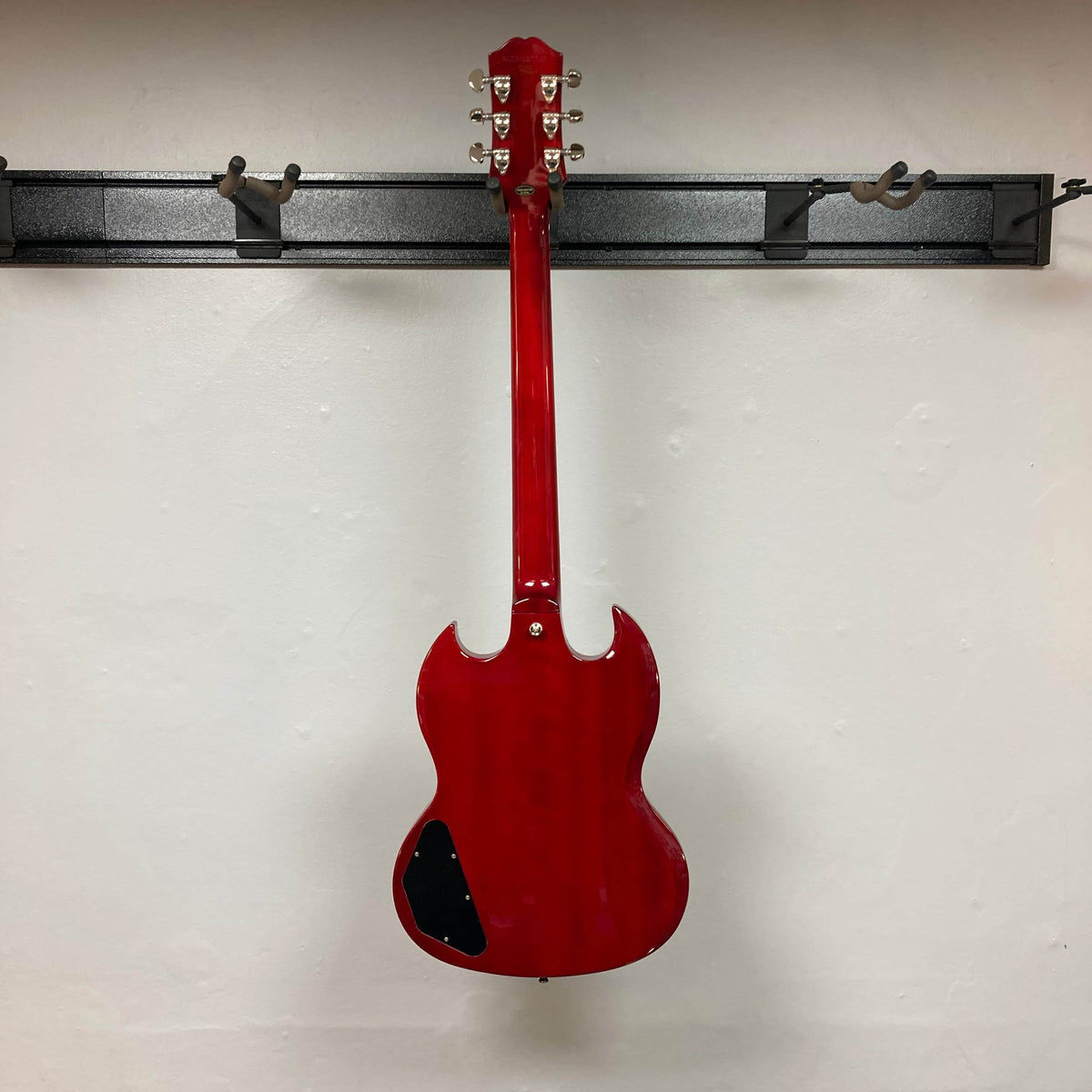 An Epiphone SG Standard Cherry w/Gigbag by Epiphone hangs on a black wall-mounted rack with its back facing outward, showcased against a white wall.