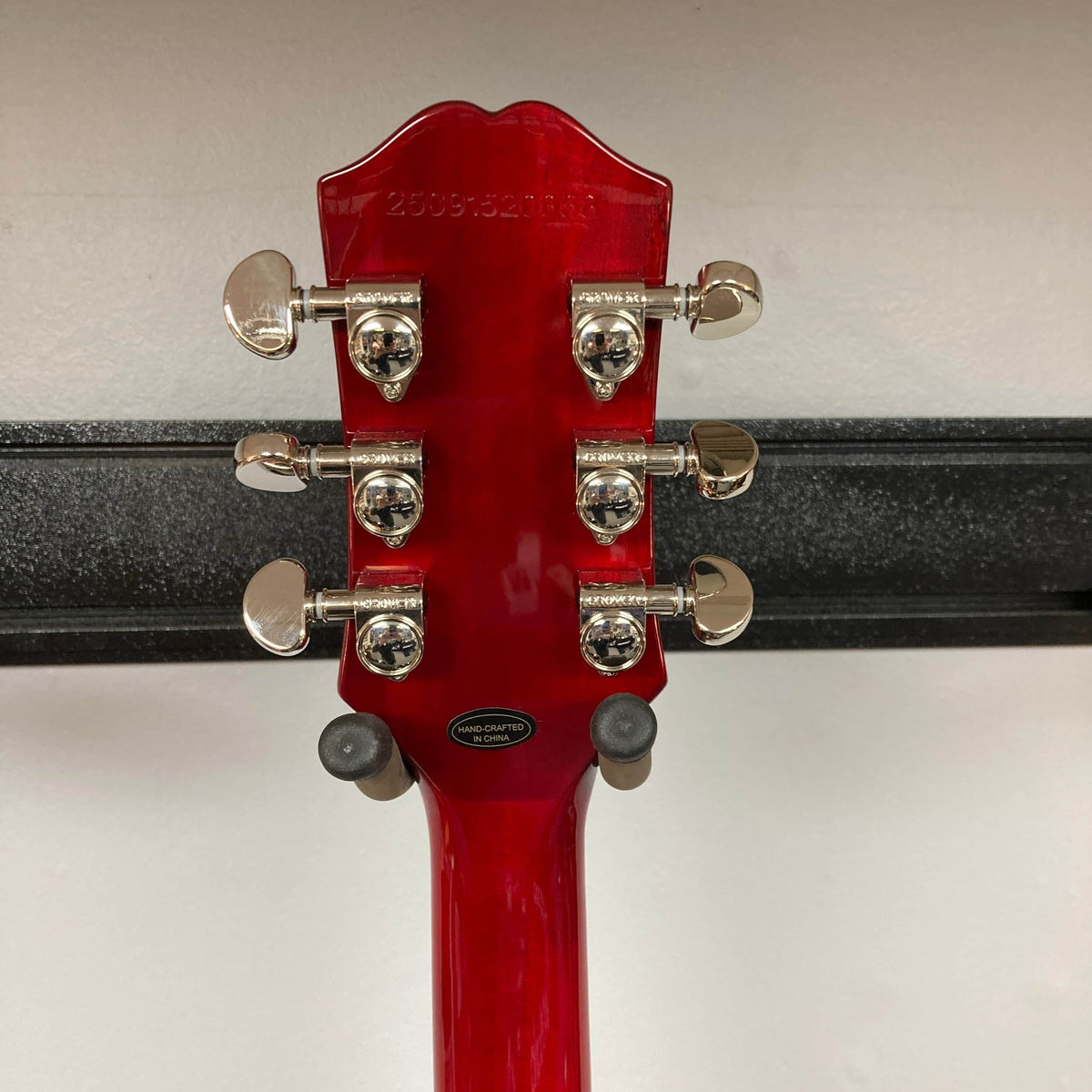 Close-up of the back of an Epiphone SG Standard Cherry w/Gigbag headstock by Epiphone, featuring six silver tuning pegs and a serial number, displayed on a black wall-mounted hanger.