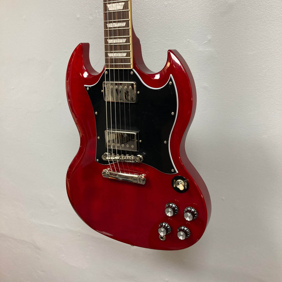 An Epiphone SG Standard Cherry w/Gigbag electric guitar by Epiphone, featuring a mahogany body, black pickguard, six strings, and four control knobs, is displayed hanging against a plain light-colored wall.